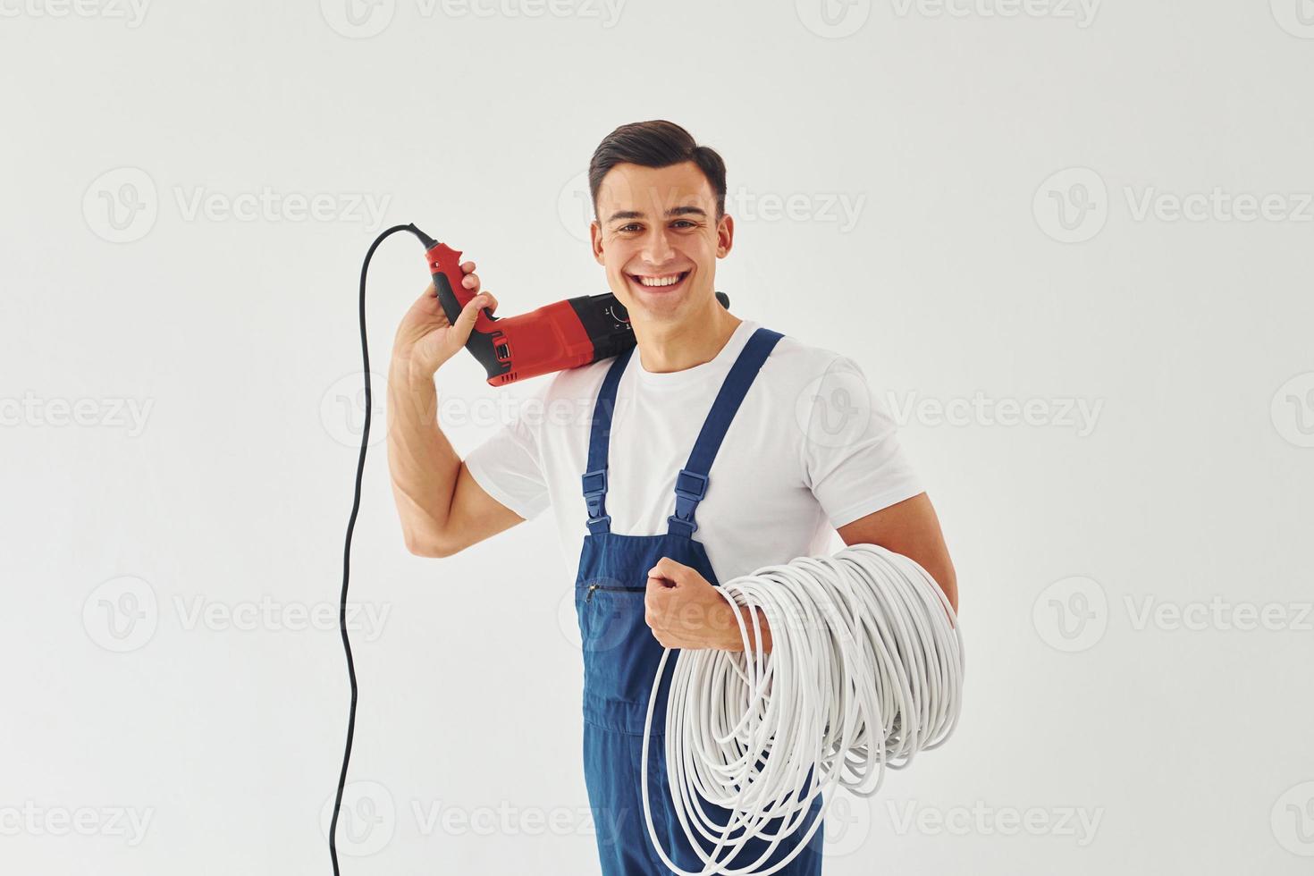 con taladro y cables en las manos. trabajador de sexo masculino en uniforme azul de pie dentro del estudio contra el fondo blanco foto