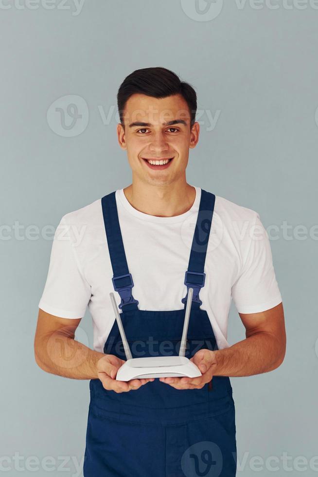 Holds wifi router. Male worker in blue uniform standing inside of studio against white background photo