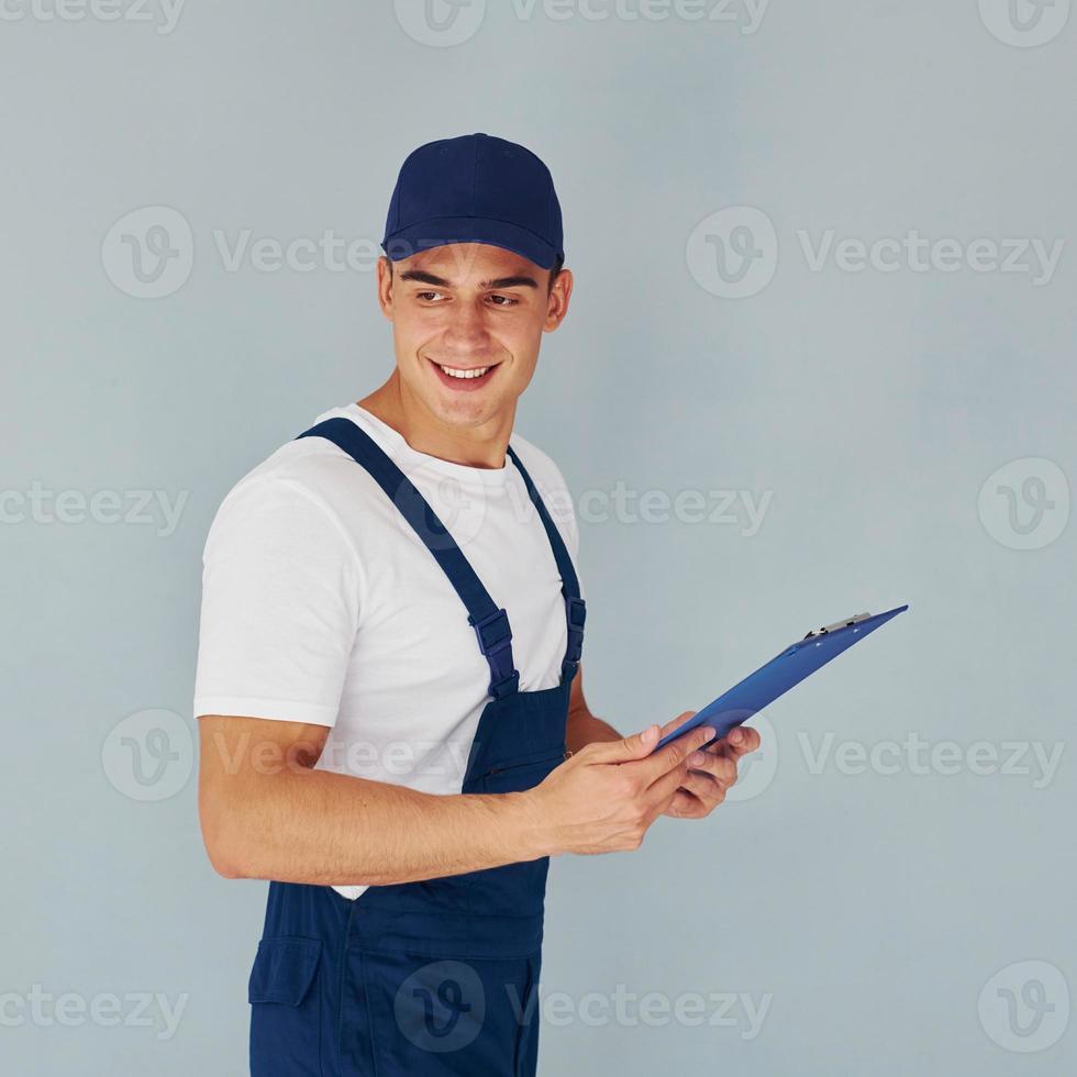 In cap and with notepad. Male worker in blue uniform standing inside of studio against white background photo