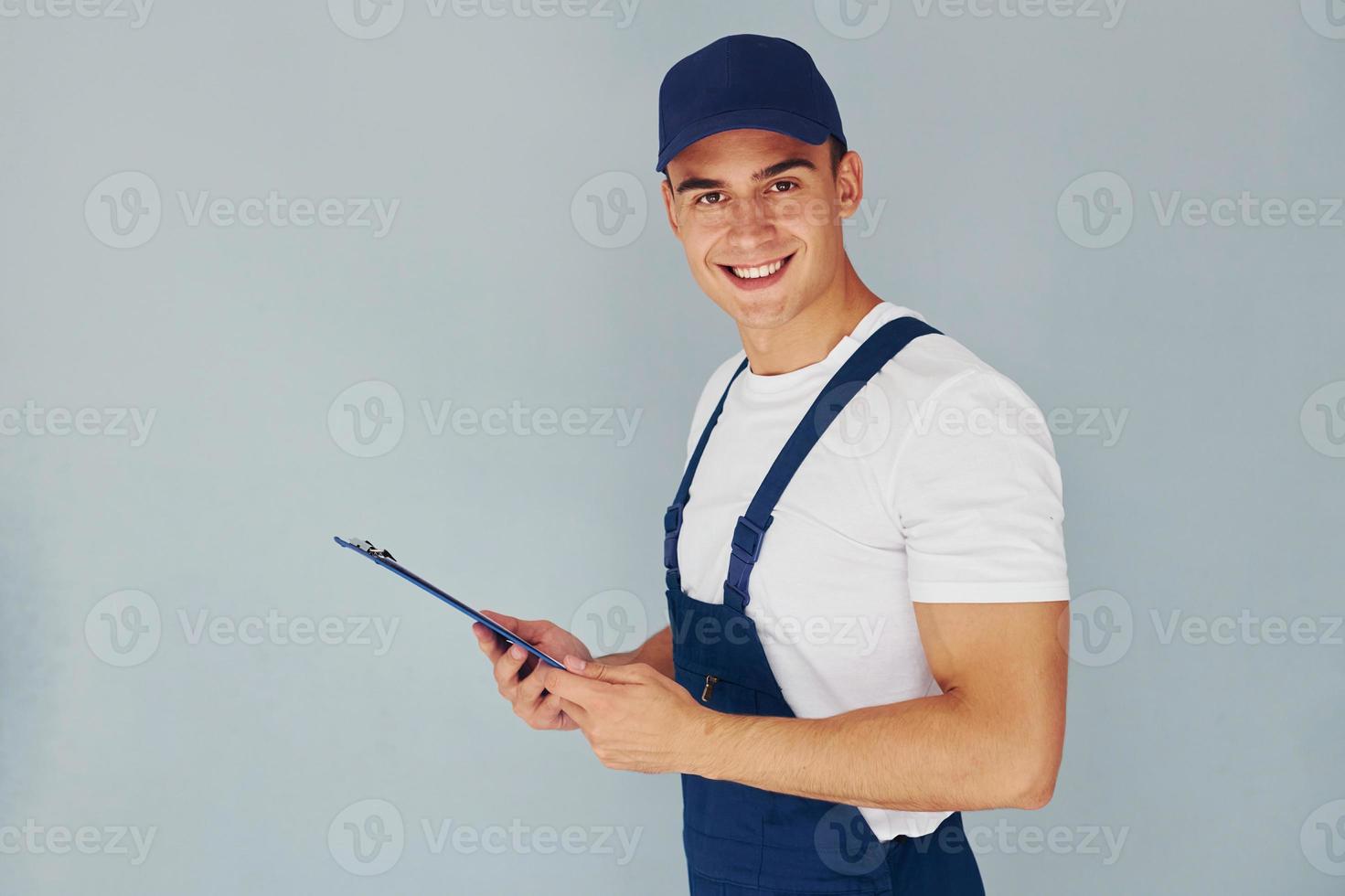 en gorra y con bloc de notas. trabajador de sexo masculino en uniforme azul de pie dentro del estudio contra el fondo blanco foto