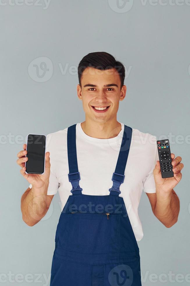 TV remote controller and smartphone. Male worker in blue uniform standing inside of studio against white background photo