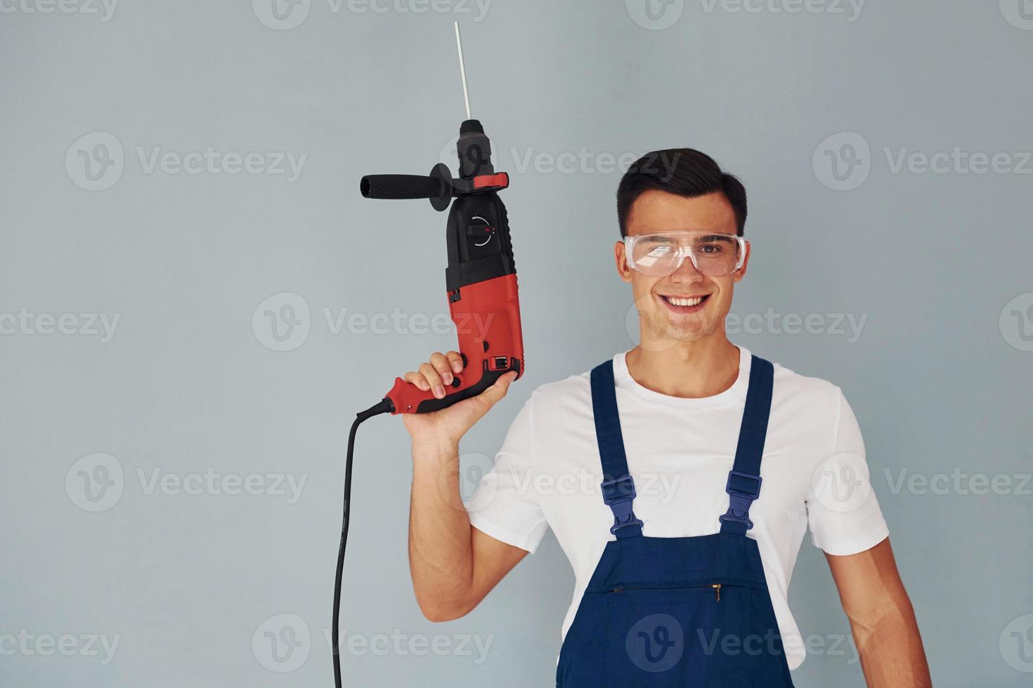 In protective eyewear and with drill in hands. Male worker in blue uniform standing inside of studio against white background photo