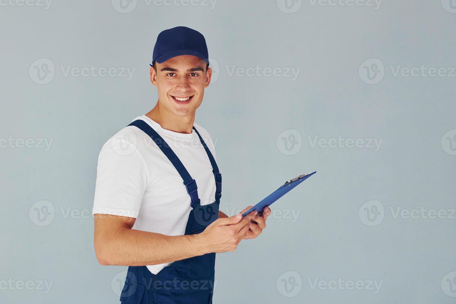 In cap and with notepad. Male worker in blue uniform standing inside of studio against white background photo