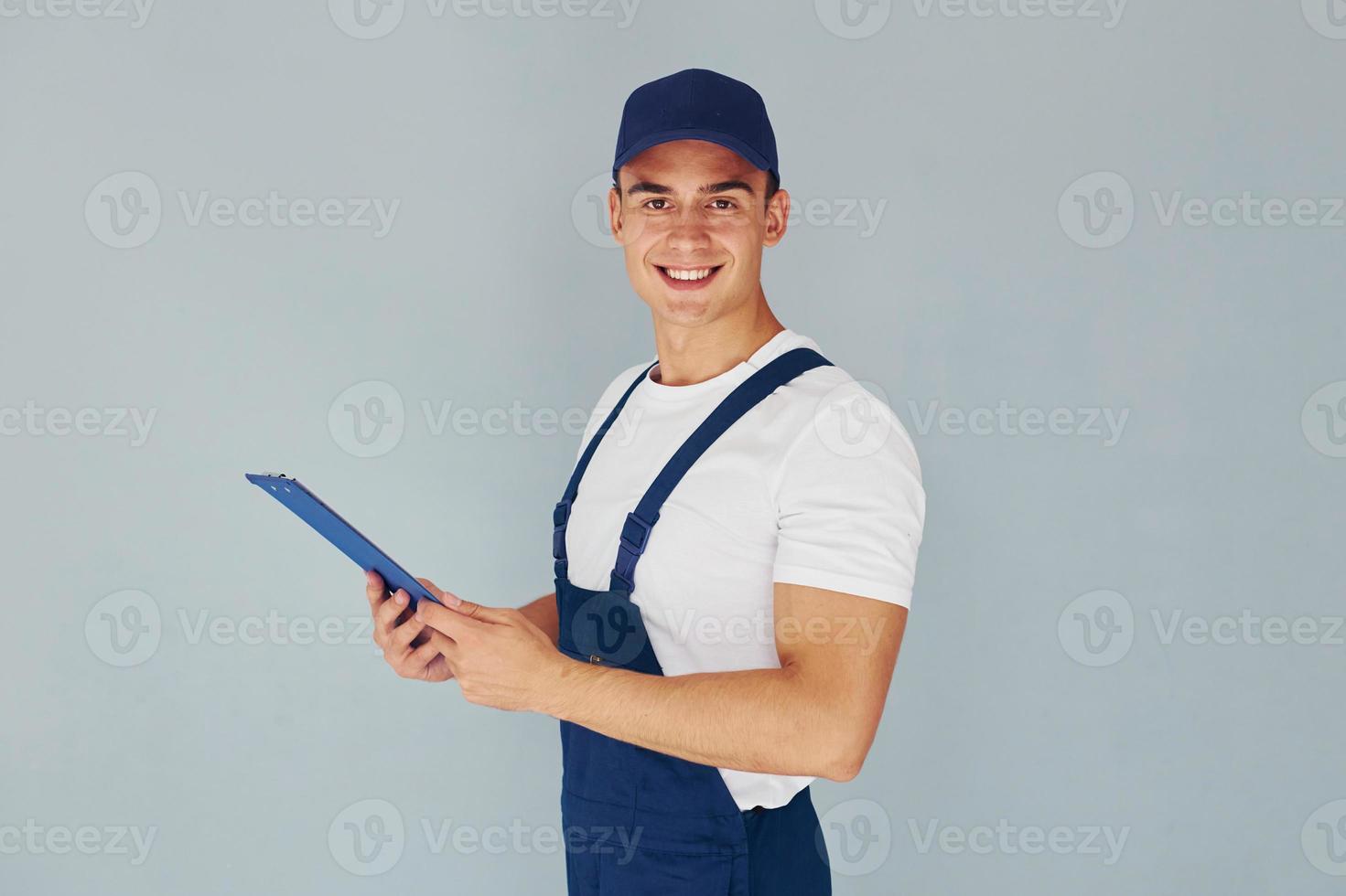 en gorra y con bloc de notas. trabajador de sexo masculino en uniforme azul de pie dentro del estudio contra el fondo blanco foto