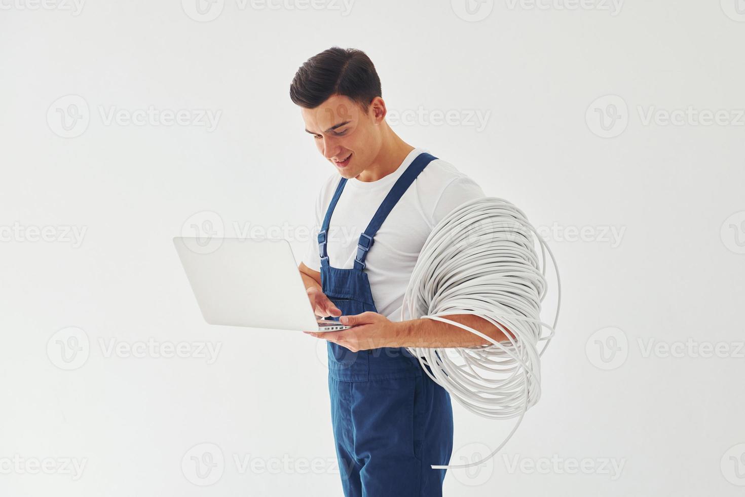 Uses laptop. Male worker in blue uniform standing inside of studio against white background photo