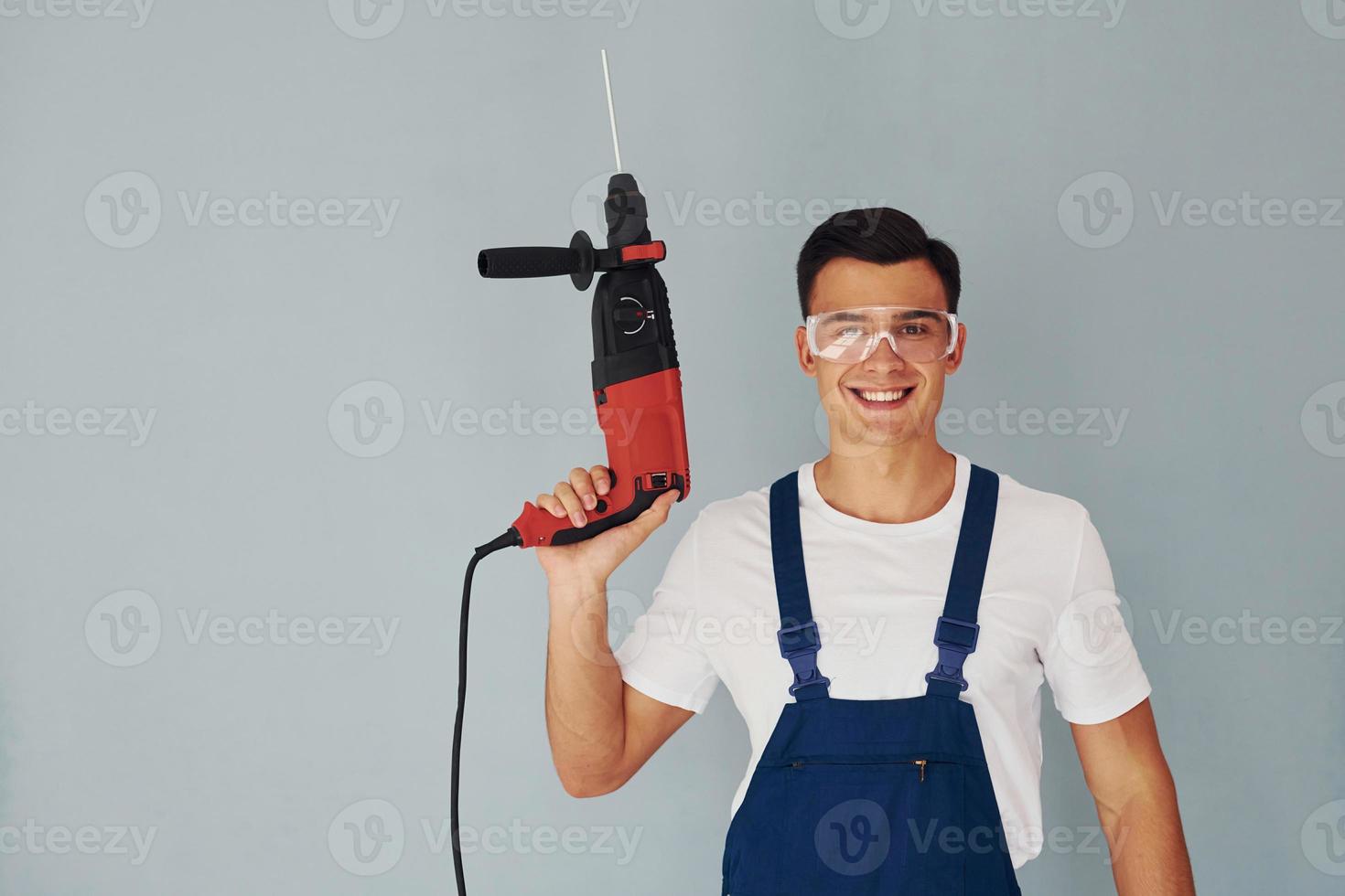 In protective eyewear and with drill in hands. Male worker in blue uniform standing inside of studio against white background photo