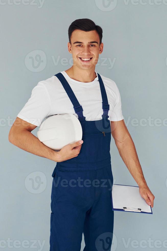 With notepad and hard hat. Male worker in blue uniform standing inside of studio against white background photo