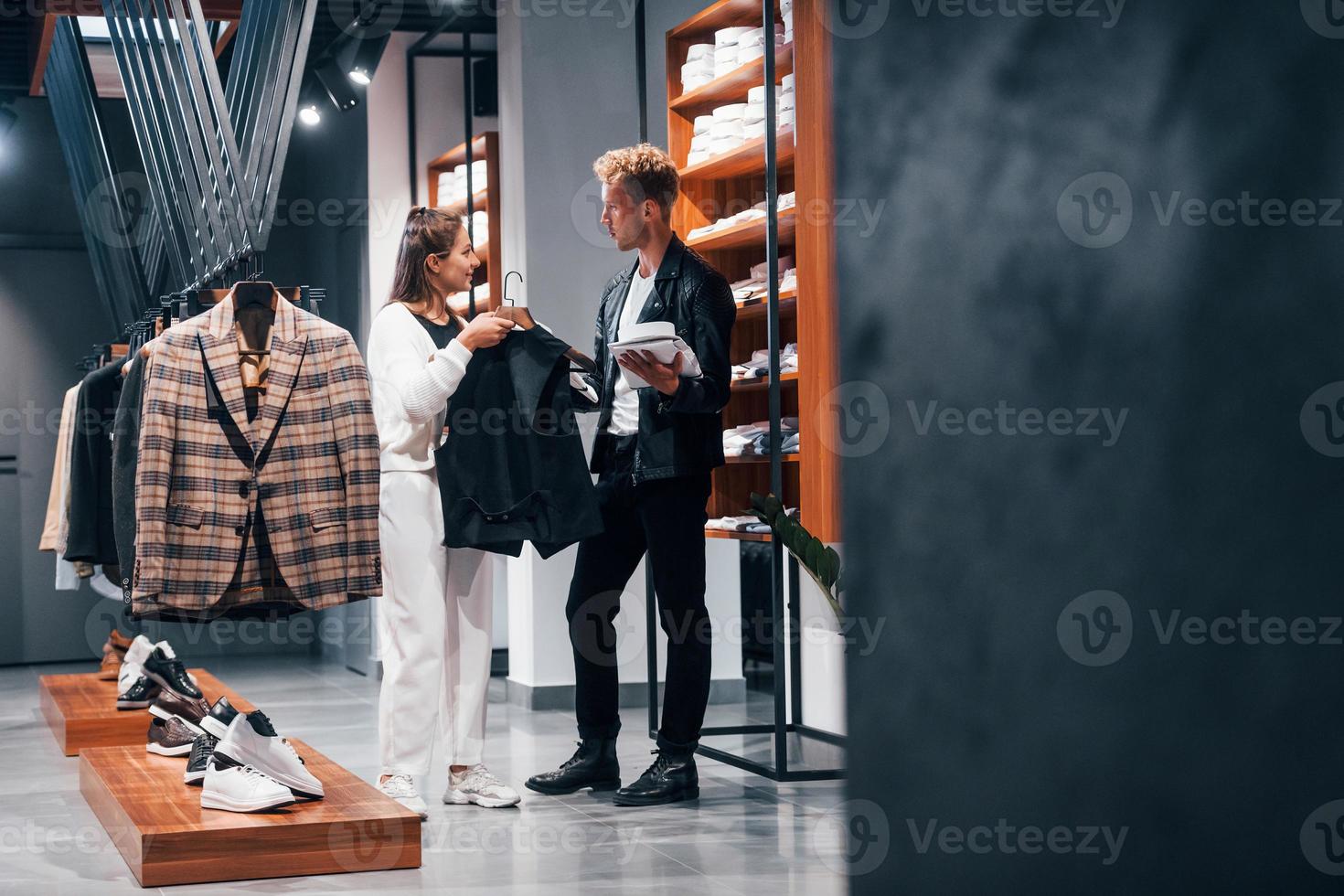 joven trabajadora ayudando a un chico a elegir ropa en una tienda moderna con ropa cara para hombres foto