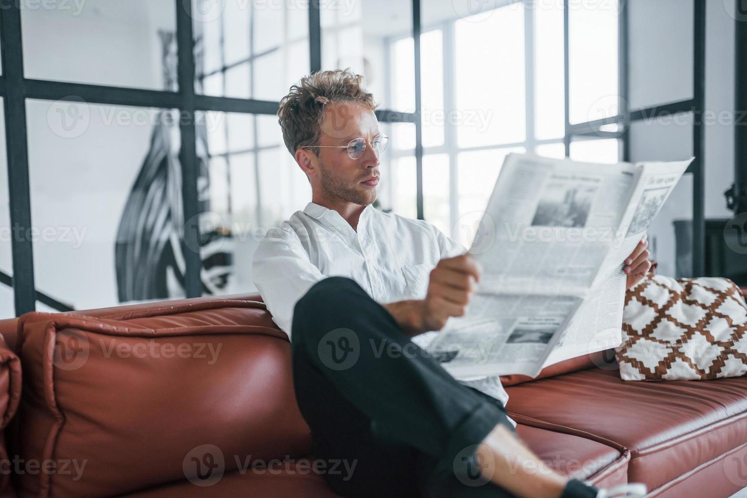 Reads newspaper. Caucasian young guy in elegant white shirt indoors at home photo
