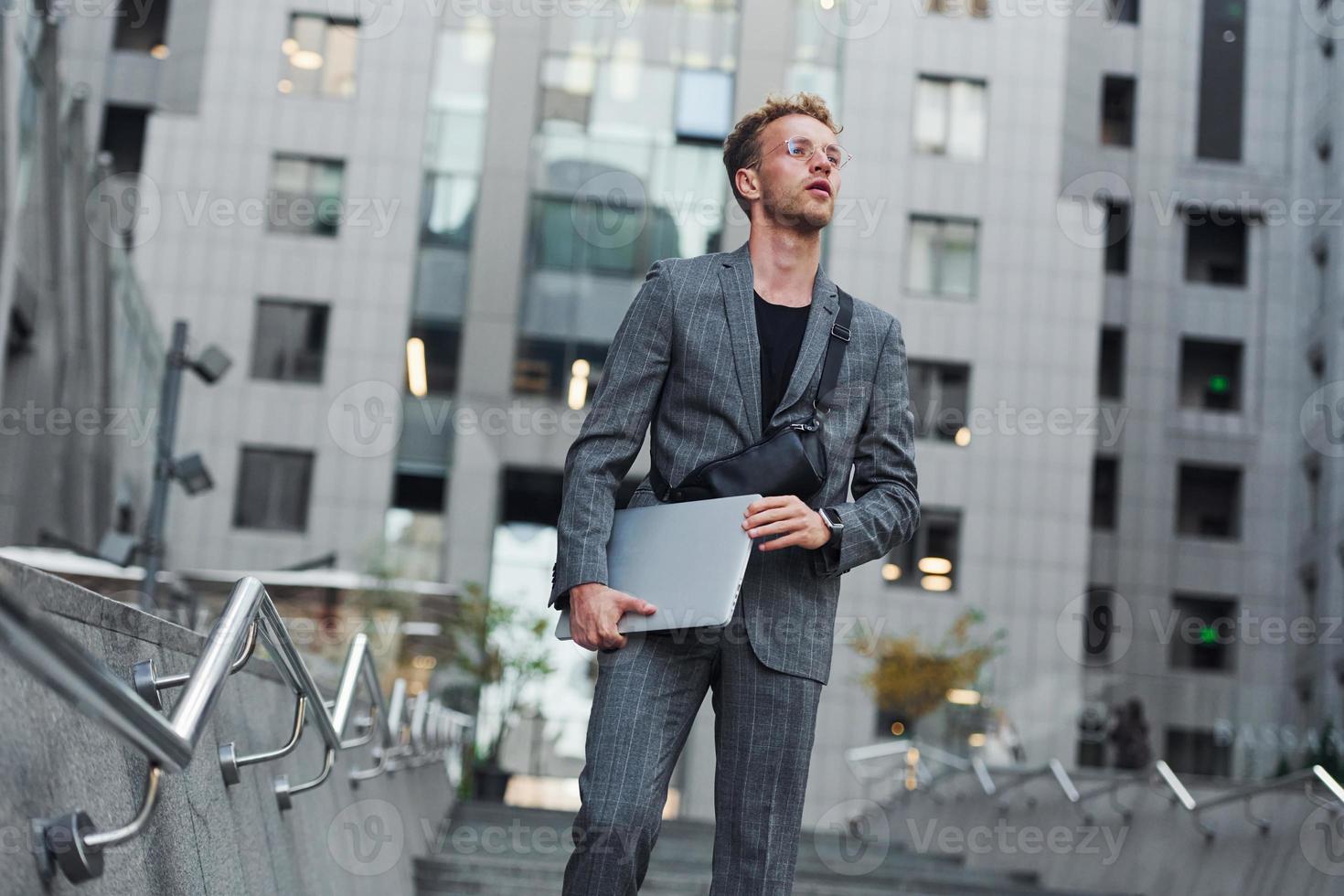 Laptop in hands. Young elegant man in good clothes is outdoors in the city at daytime photo
