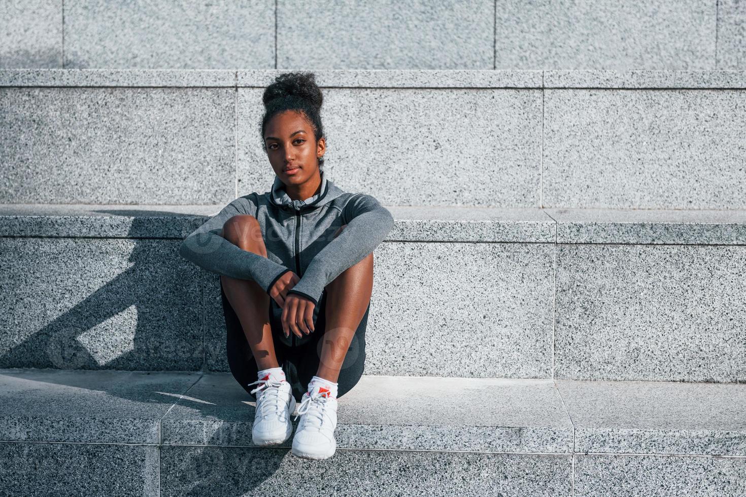 Sits on stairs. Young african american woman in sportive clothes have workout outdoors at daytime photo