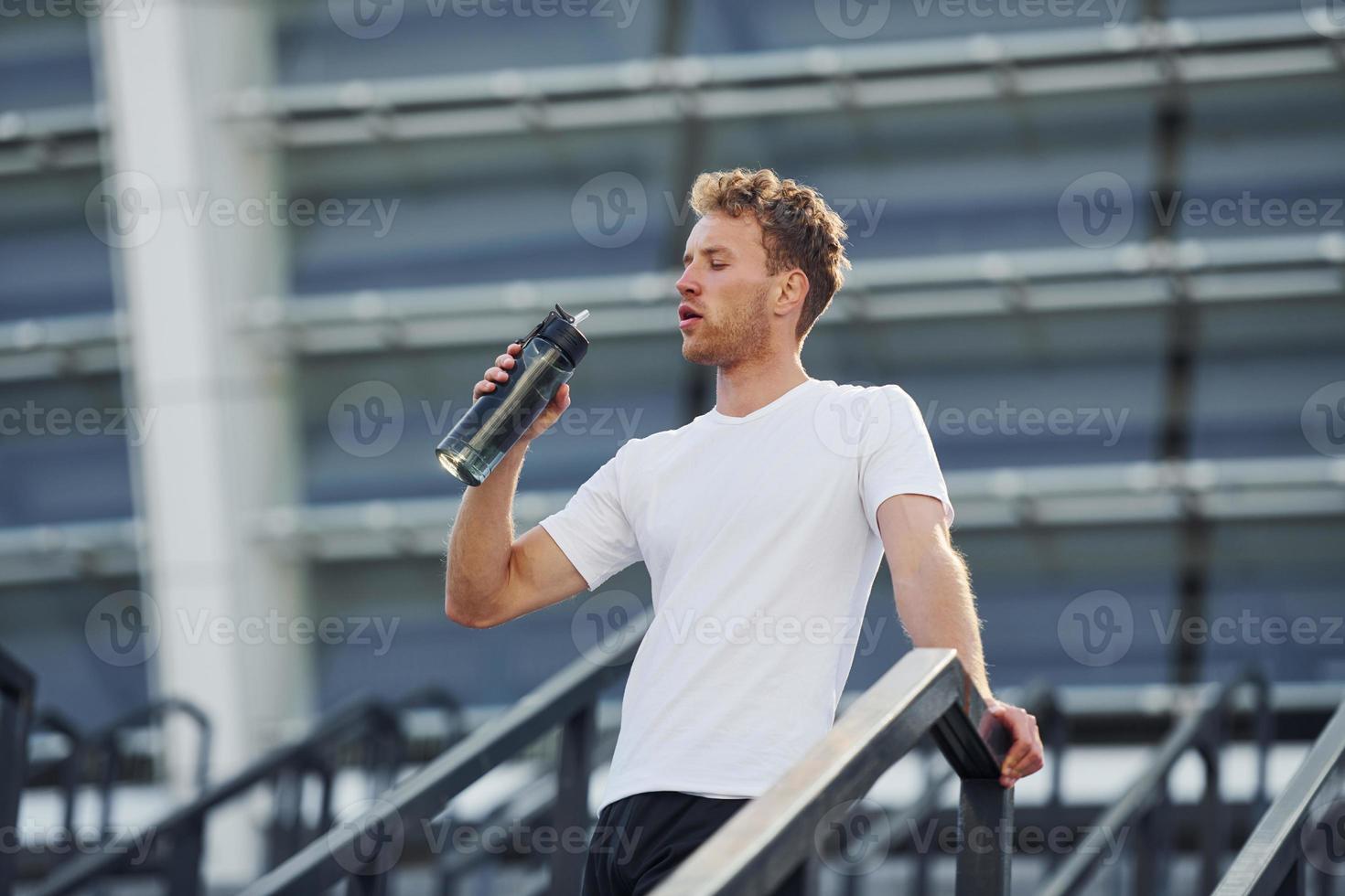Drinks water and takes a break. Young man in sportive clothes have workout outdoors at daytime photo