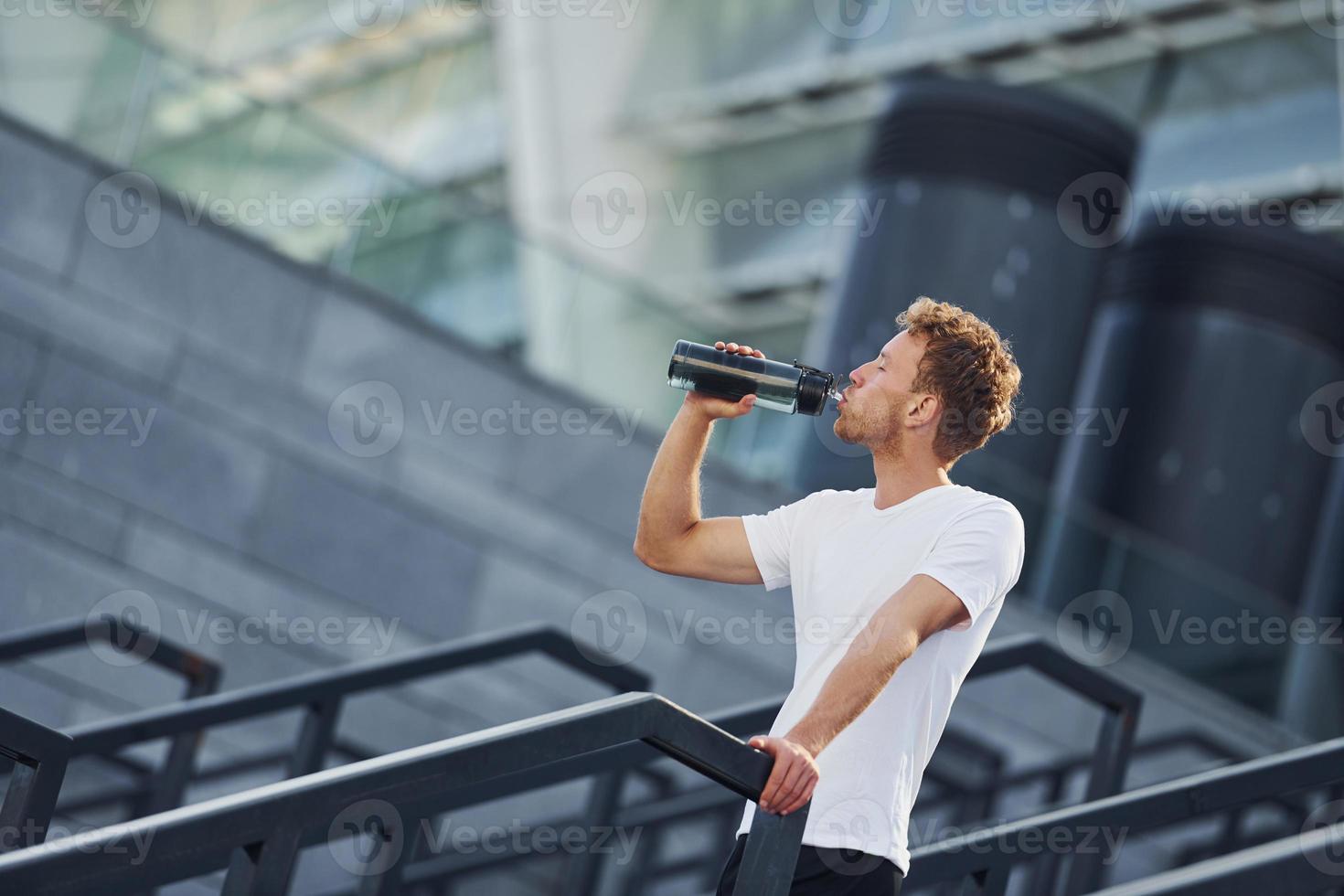 Drinks water and takes a break. Young man in sportive clothes have workout outdoors at daytime photo