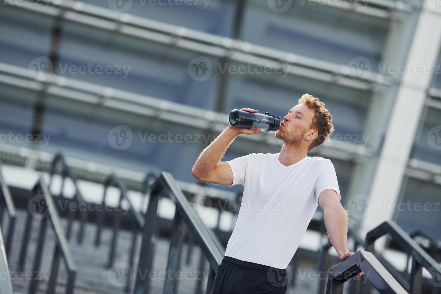 Drinks water and takes a break. Young man in sportive clothes have workout outdoors at daytime photo