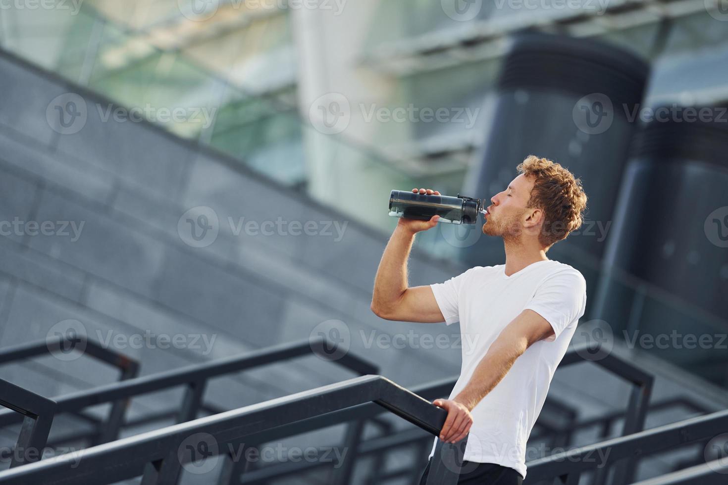 Drinks water and takes a break. Young man in sportive clothes have workout outdoors at daytime photo