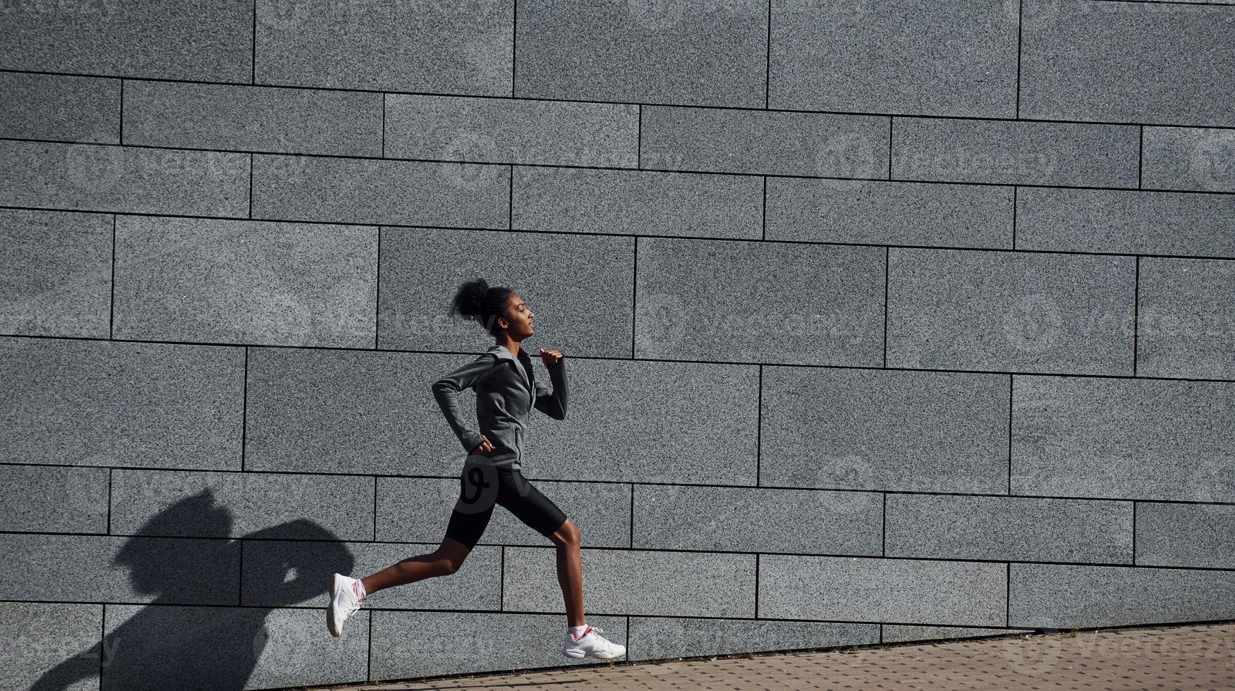 Side view of young african american woman in sportive clothes that runs near wall photo