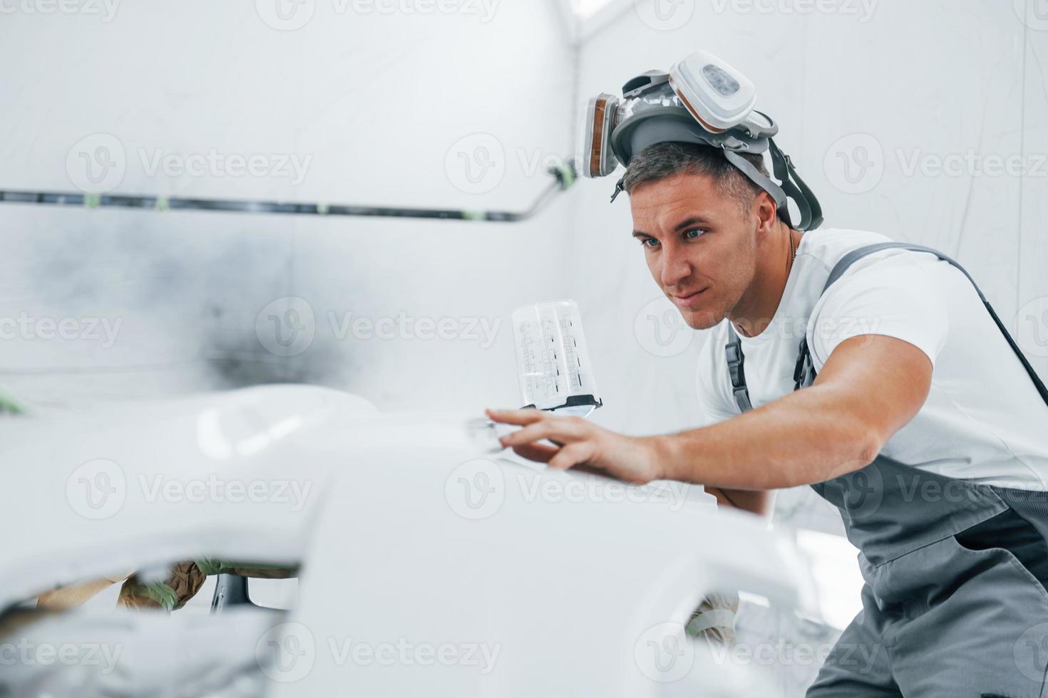 With painting gun. Caucasian automobile repairman in uniform works in garage photo