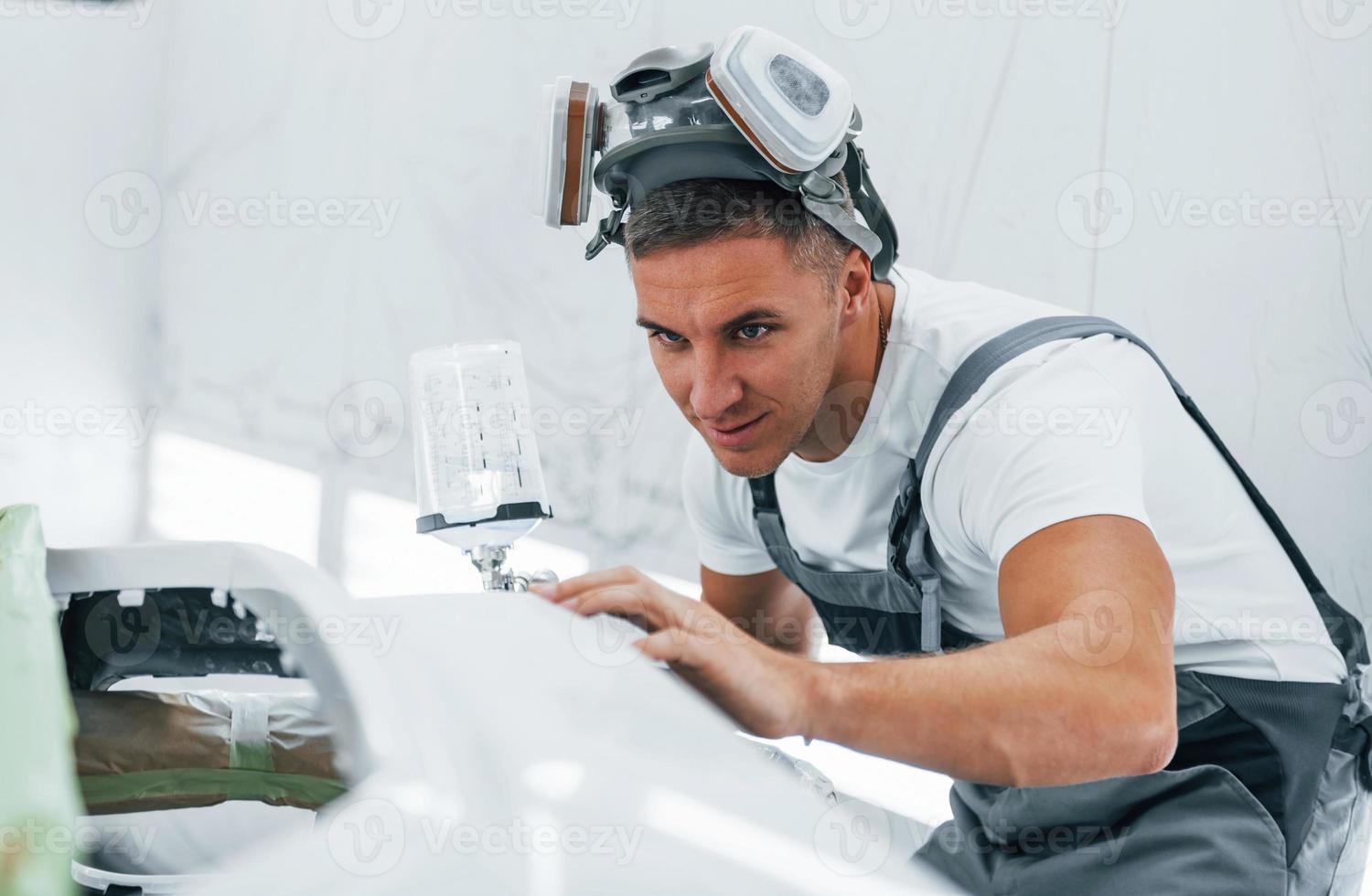 With painting gun. Caucasian automobile repairman in uniform works in garage photo