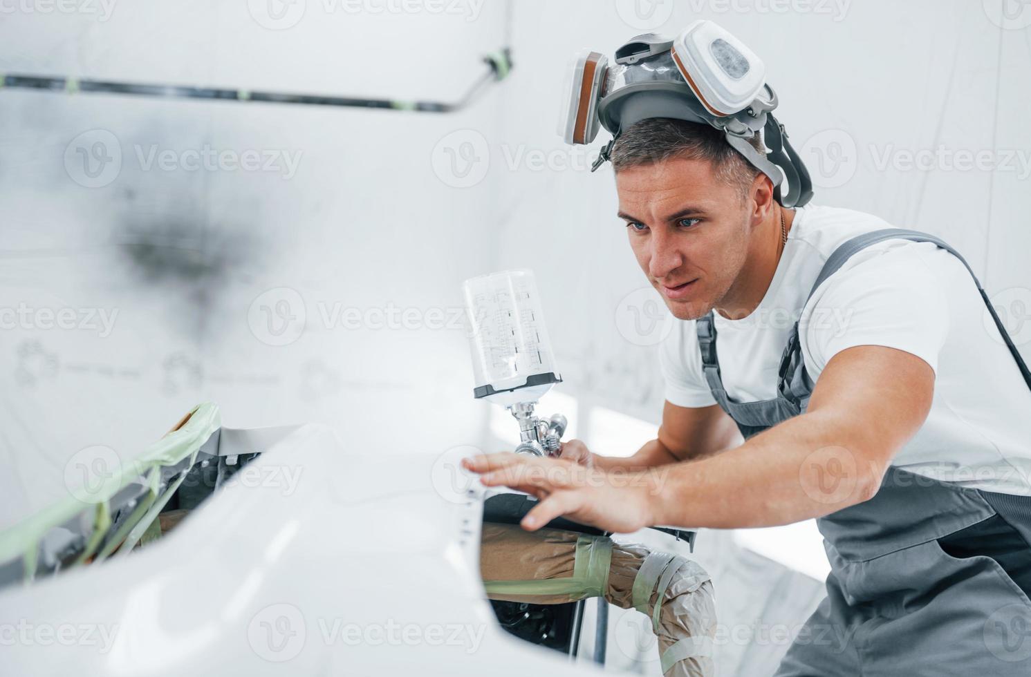 With painting gun. Caucasian automobile repairman in uniform works in garage photo