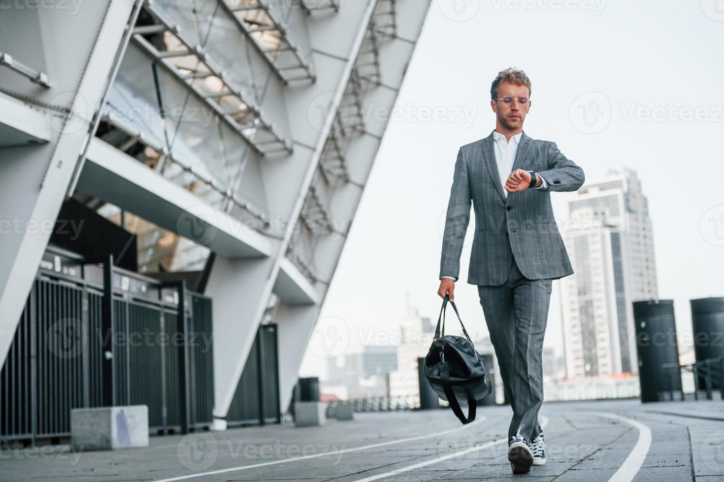 Checks time. Young businessman in grey formal wear is outdoors in the city photo