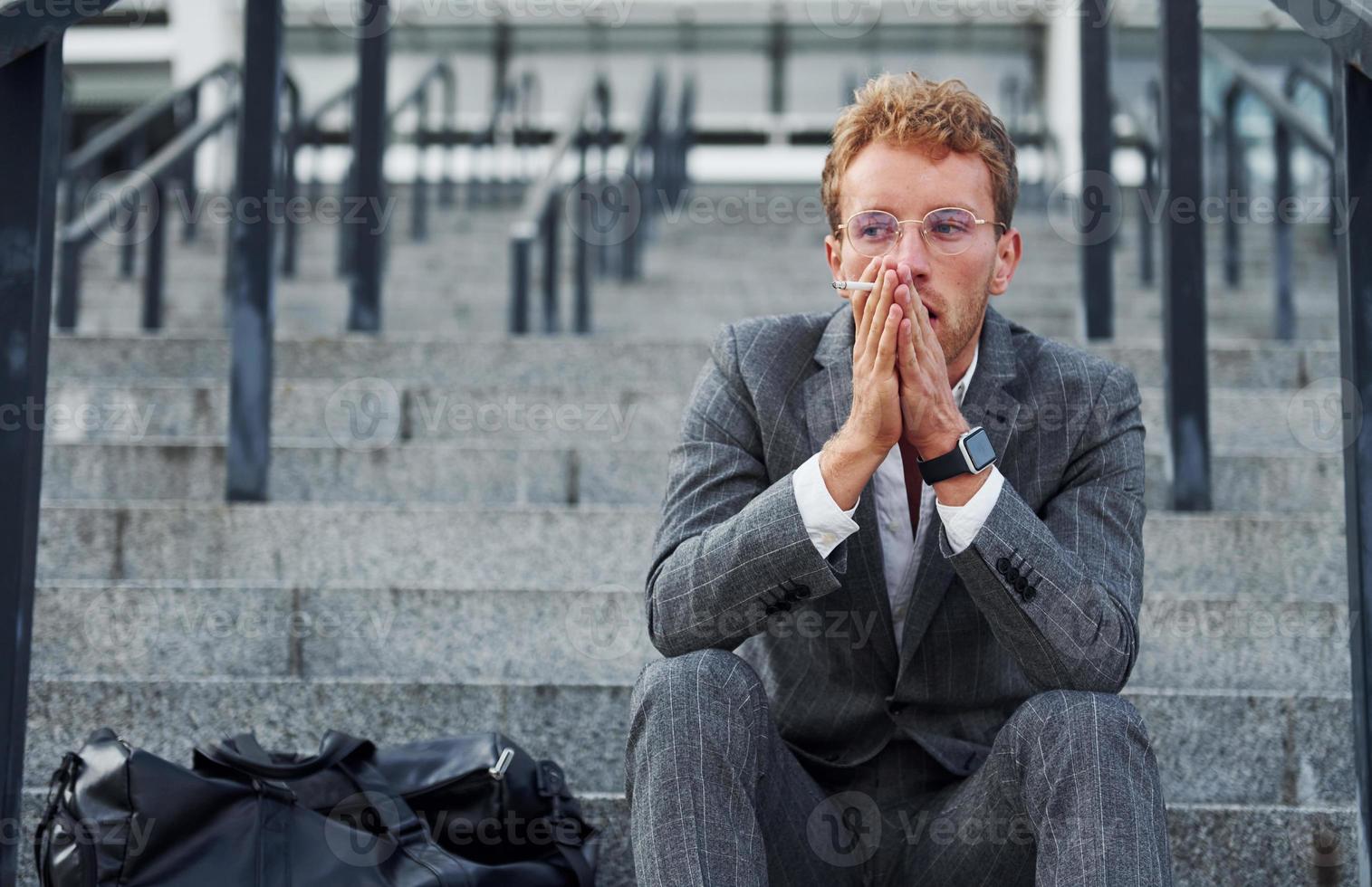 Smoking cigarette. Young businessman in grey formal wear is outdoors in the city photo