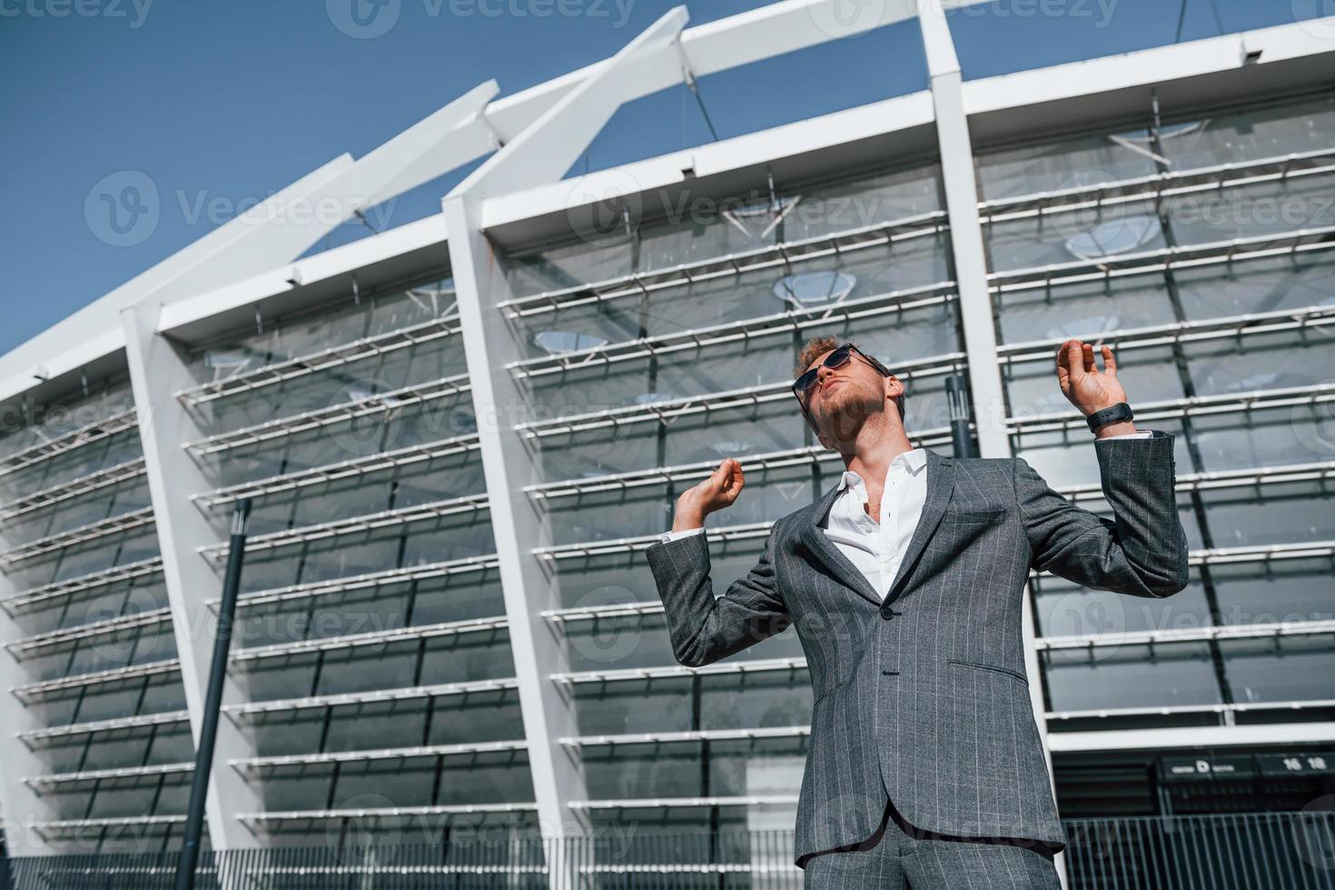 Big stadium behind. Young businessman in grey formal wear is outdoors in the city photo