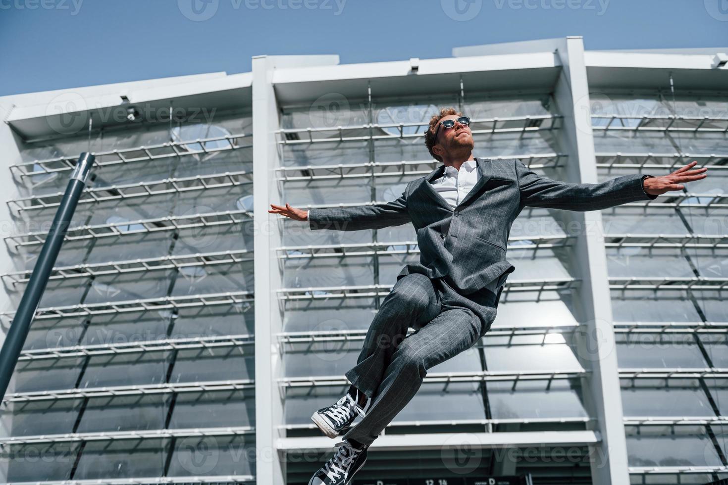 Jumping and celebrating success. Young businessman in grey formal wear is outdoors in the city photo
