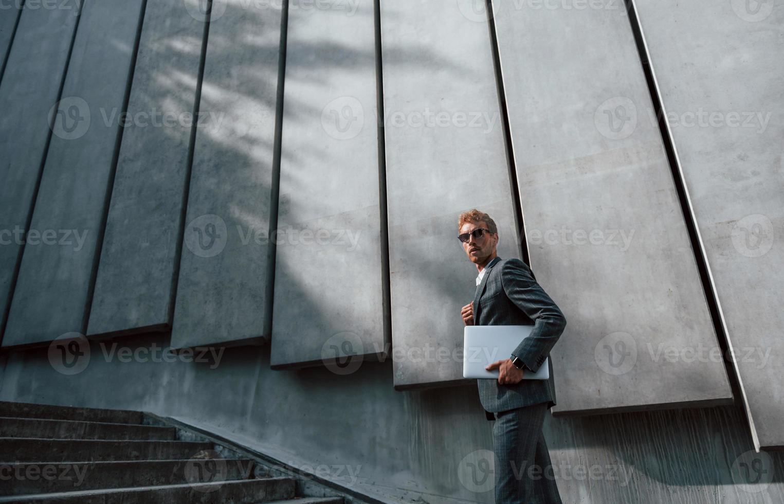 tiene la computadora portátil en las manos. un joven hombre de negocios con ropa formal gris está al aire libre en la ciudad foto
