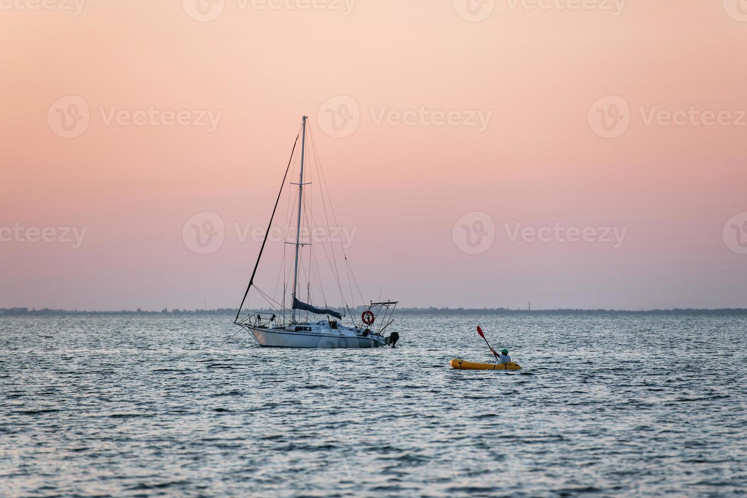 Distant view of man in the yellow colored boat near yacht that is in the sea photo