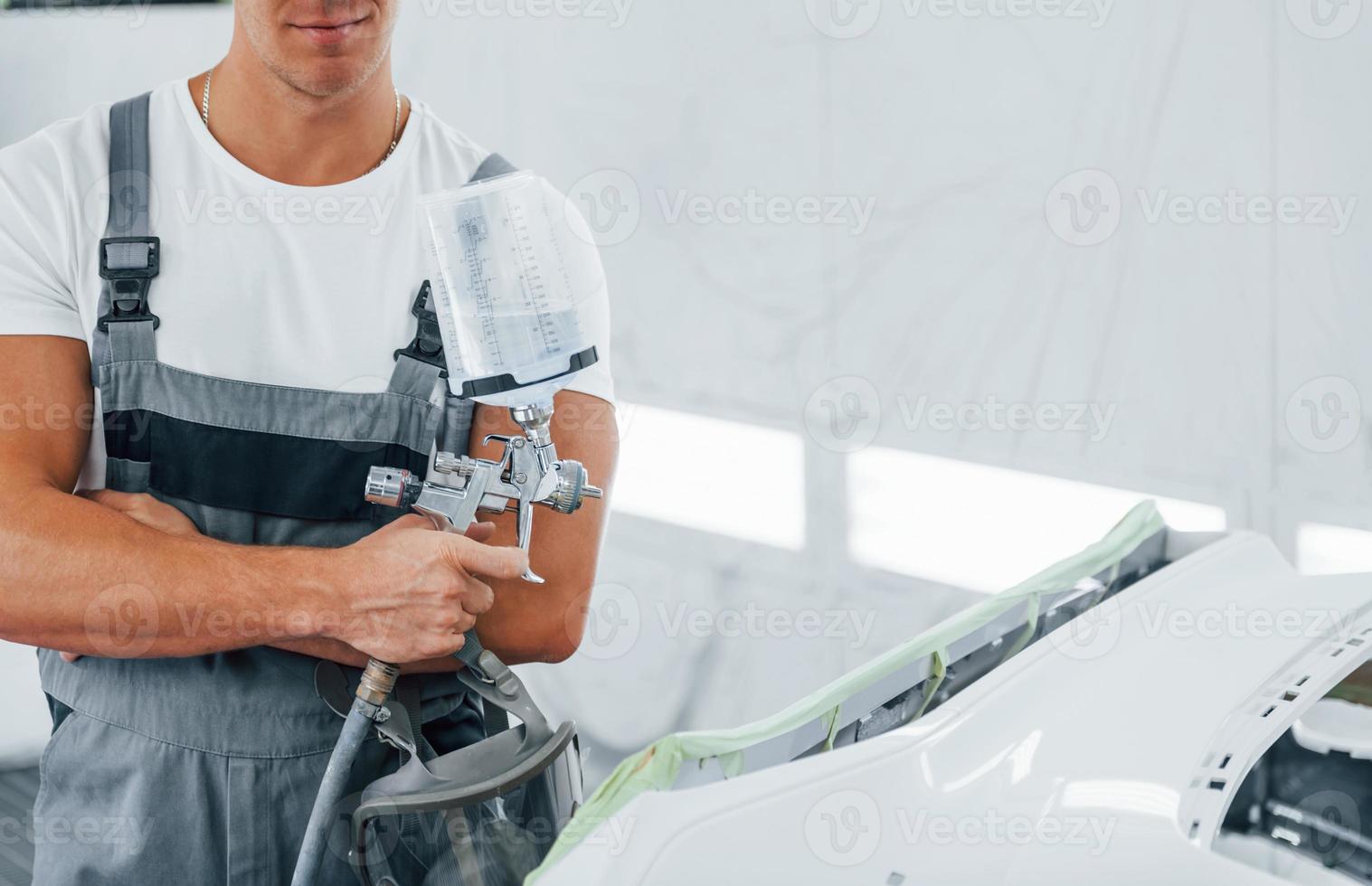 With painting gun. Caucasian automobile repairman in uniform works in garage photo