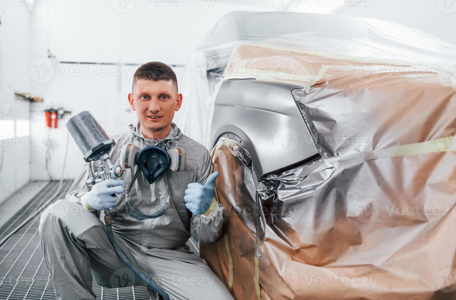 Sits near freshly painted car. Caucasian automobile repairman in uniform works in garage photo