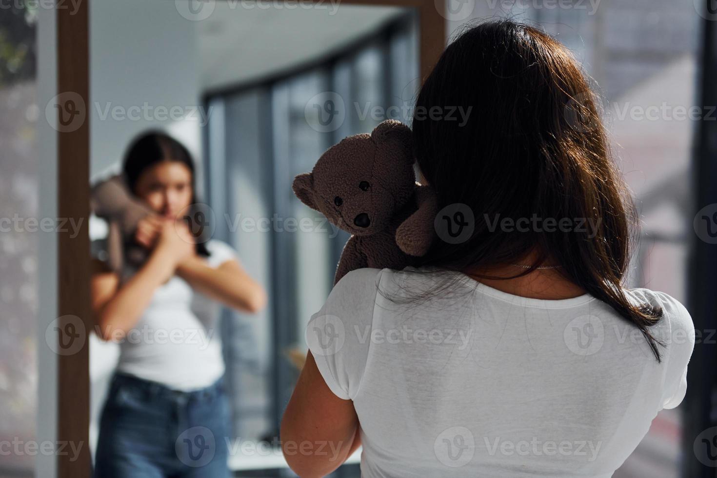 Young woman holds teddy bear and looks at herself into the mirror photo