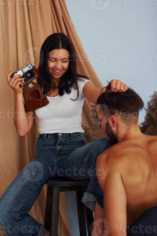 Girl with retro camera sits on the chair and trying to make photoshoot to the guy that is shirtless photo