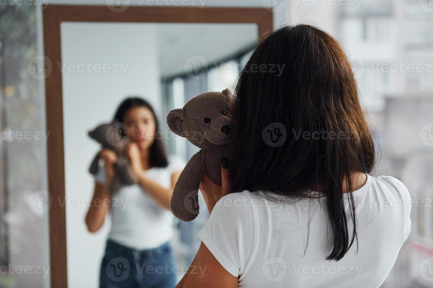 Young woman holds teddy bear and looks at herself into the mirror photo