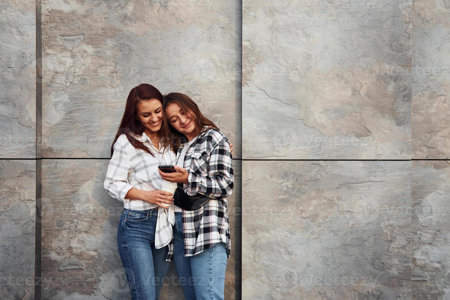 Holding drink and phone. Beautiful cheerful friends or lesbian couple together near wall outdoors at daytime photo