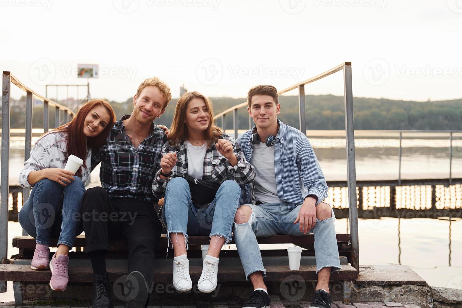 Sitting near lake and embracing each other. Group of young cheerful friends that is outdoors having fun together photo