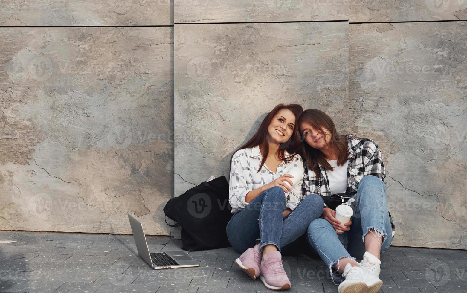 With cup of coffee. Beautiful cheerful friends or lesbian couple together near wall outdoors at daytime photo