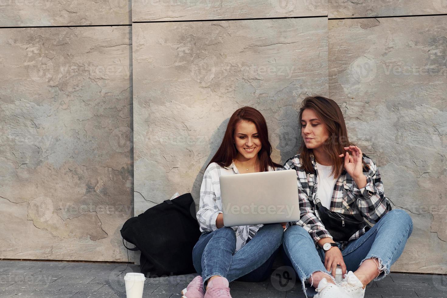 Using laptop. Beautiful cheerful friends or lesbian couple together near wall outdoors at daytime photo