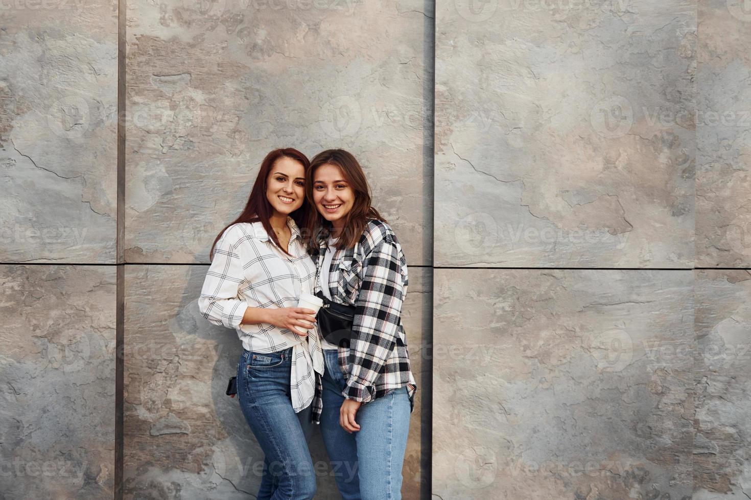 With cup of coffee. Beautiful cheerful friends or lesbian couple together near wall outdoors at daytime photo