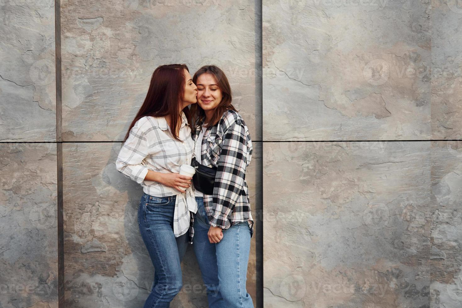 With cup of coffee. Beautiful cheerful friends or lesbian couple together near wall outdoors at daytime photo