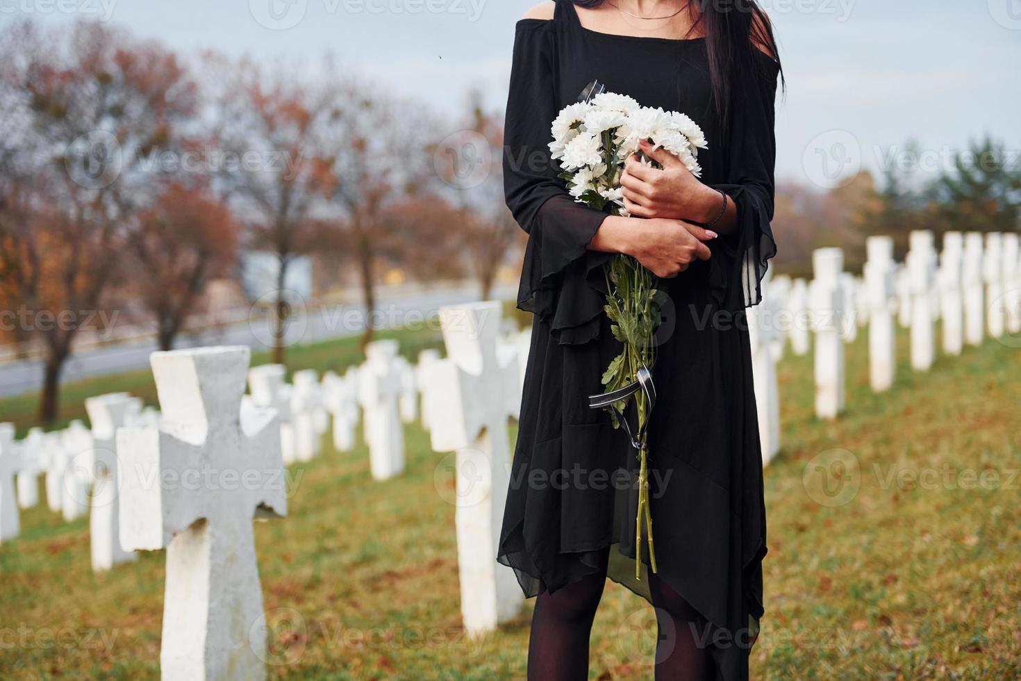 With flowers in hands. Young woman in black clothes visiting cemetery with many white crosses. Conception of funeral and death photo