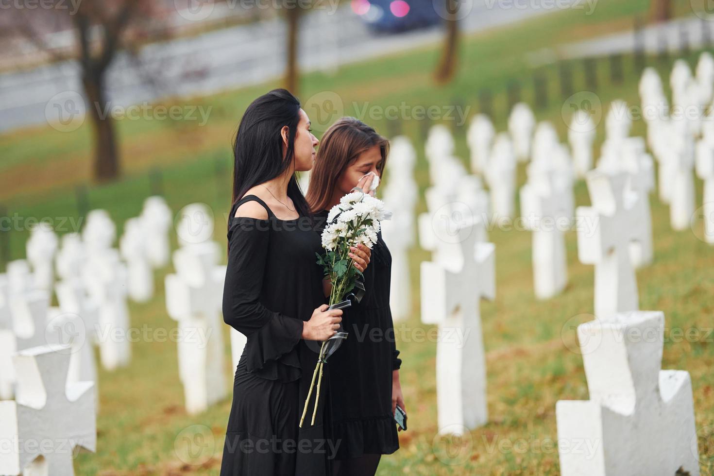 sostiene flores. dos mujeres jóvenes vestidas de negro visitando el cementerio con muchas cruces blancas. concepción del funeral y la muerte foto