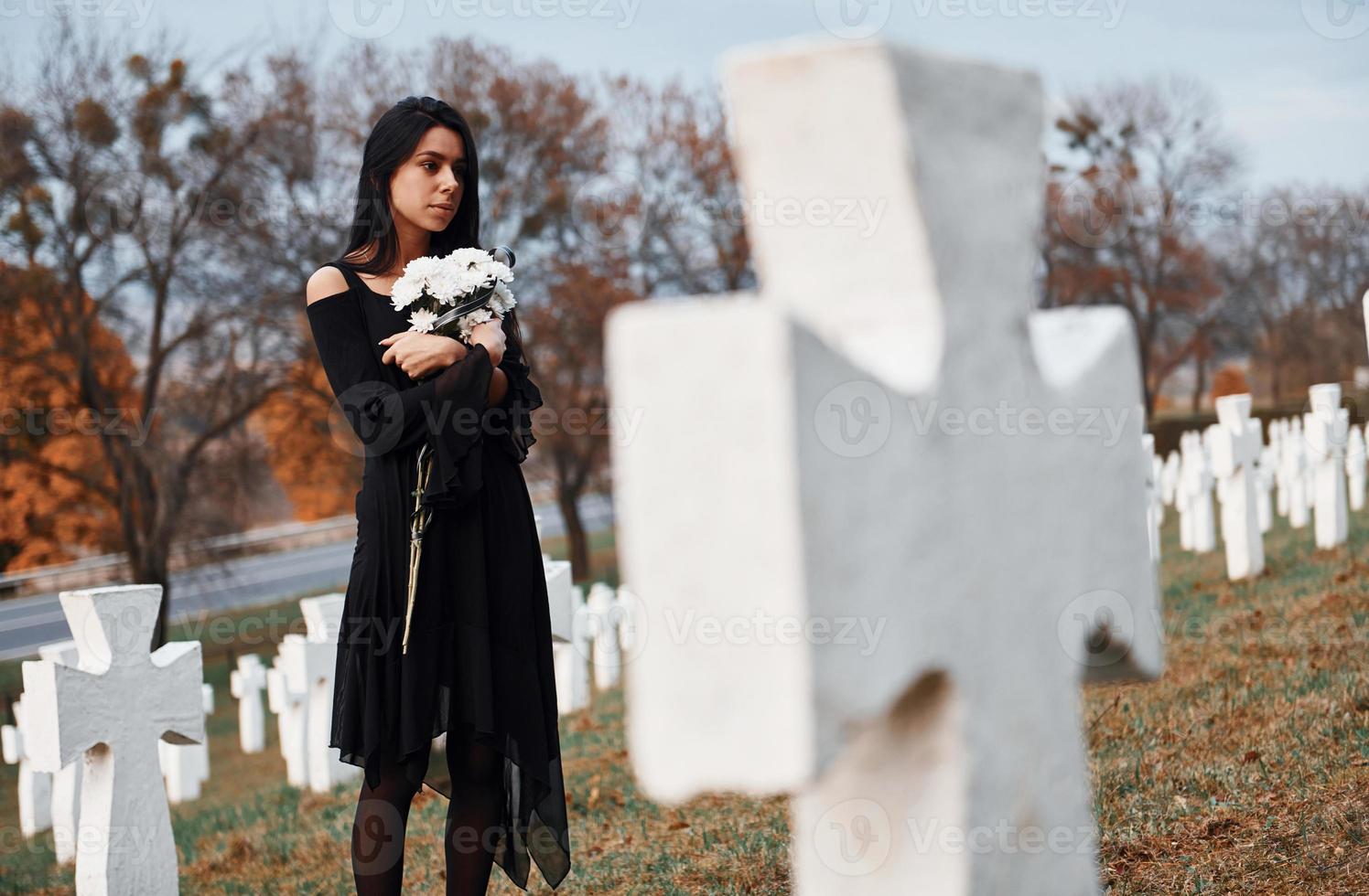 con flores en las manos. joven vestida de negro visitando el cementerio con muchas cruces blancas. concepción del funeral y la muerte foto