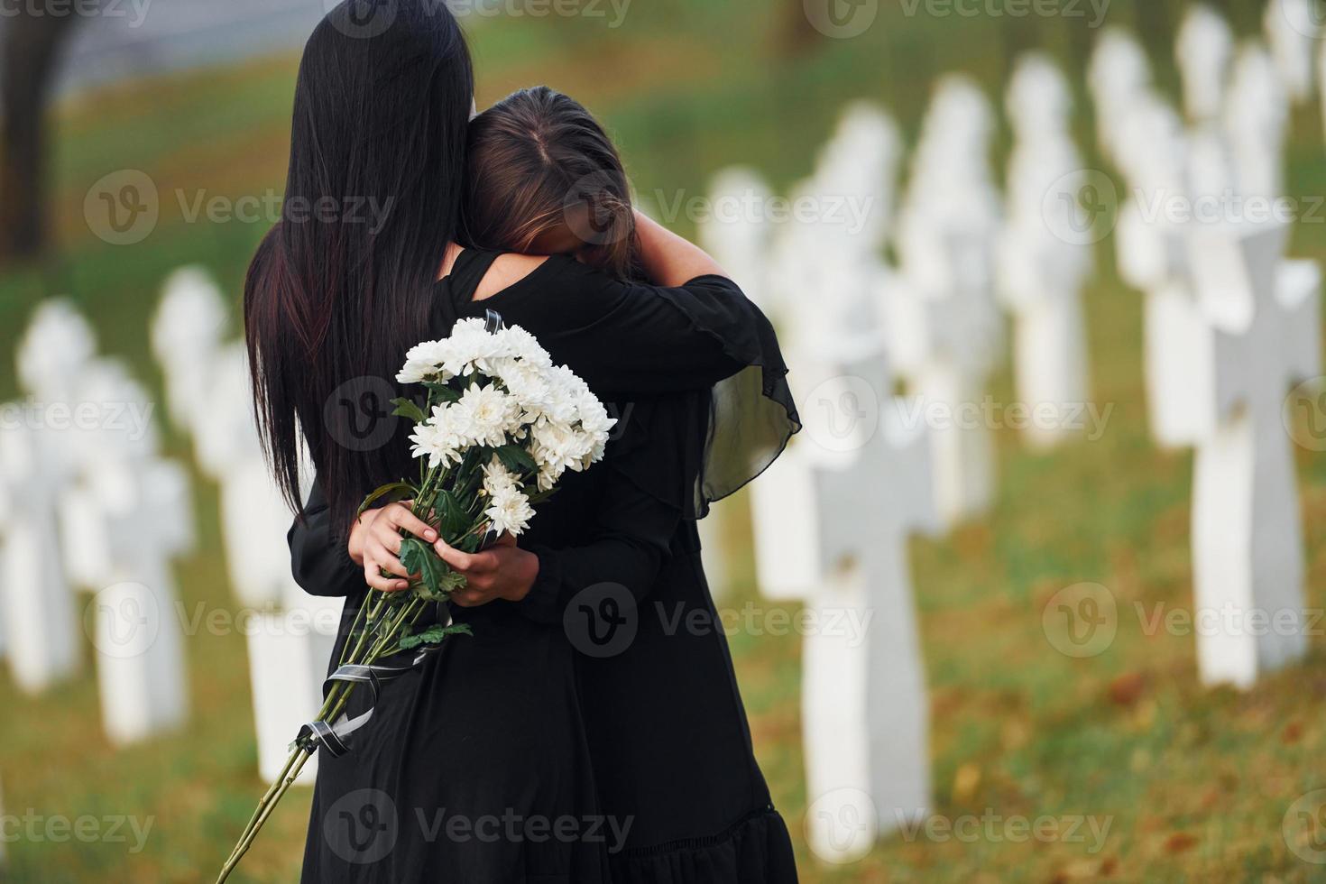 abrazándose y llorando. dos mujeres jóvenes vestidas de negro visitando el cementerio con muchas cruces blancas. concepción del funeral y la muerte foto