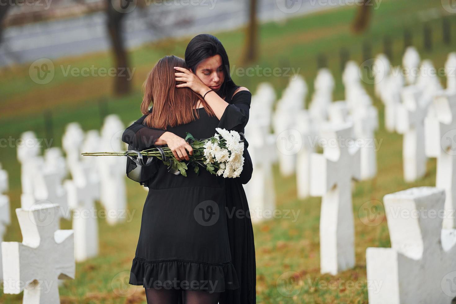 abrazándose y llorando. dos mujeres jóvenes vestidas de negro visitando el cementerio con muchas cruces blancas. concepción del funeral y la muerte foto