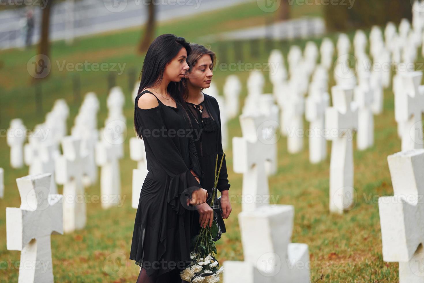 sostiene flores. dos mujeres jóvenes vestidas de negro visitando el cementerio con muchas cruces blancas. concepción del funeral y la muerte foto