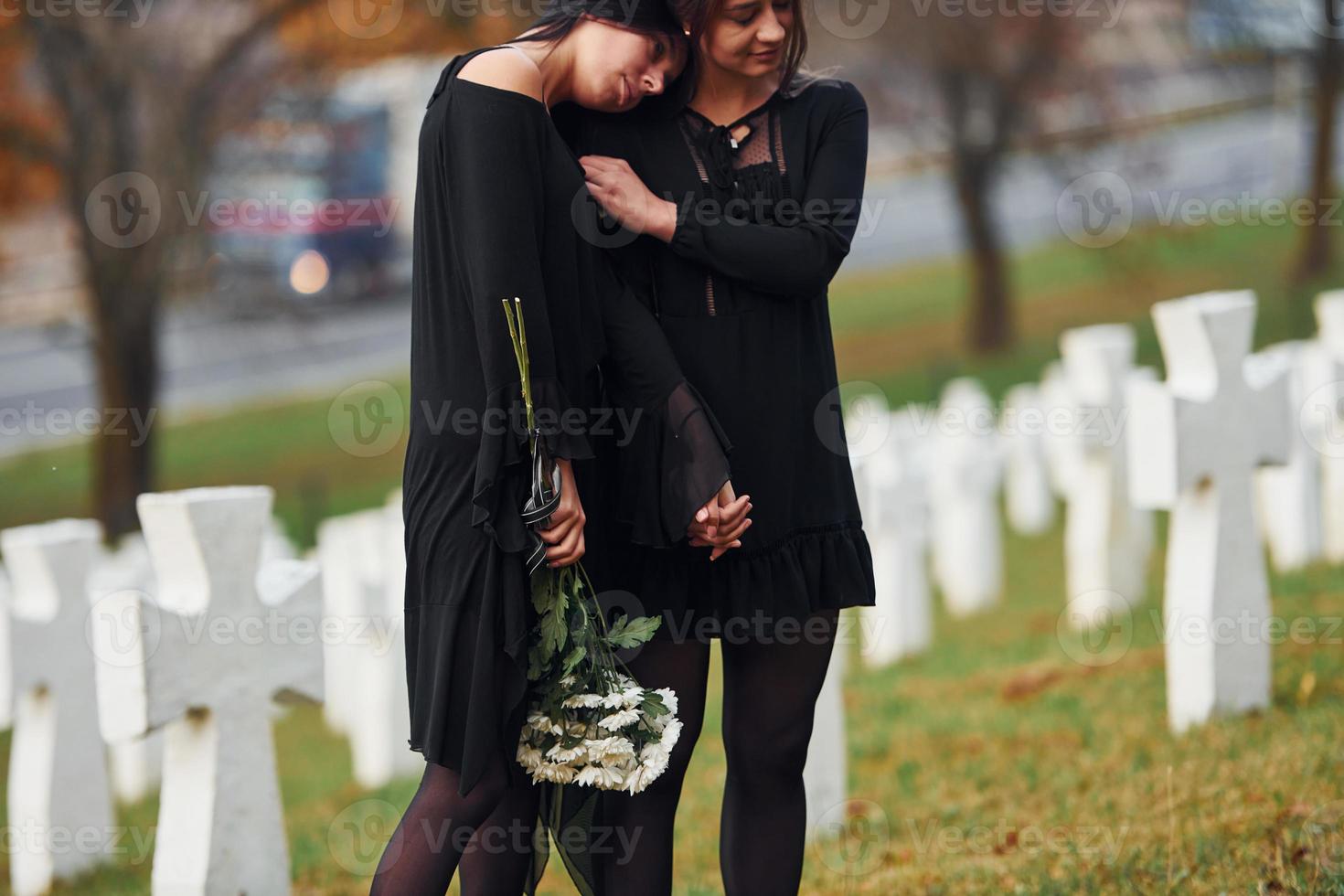 Embracing each other and crying. Two young women in black clothes visiting cemetery with many white crosses. Conception of funeral and death photo