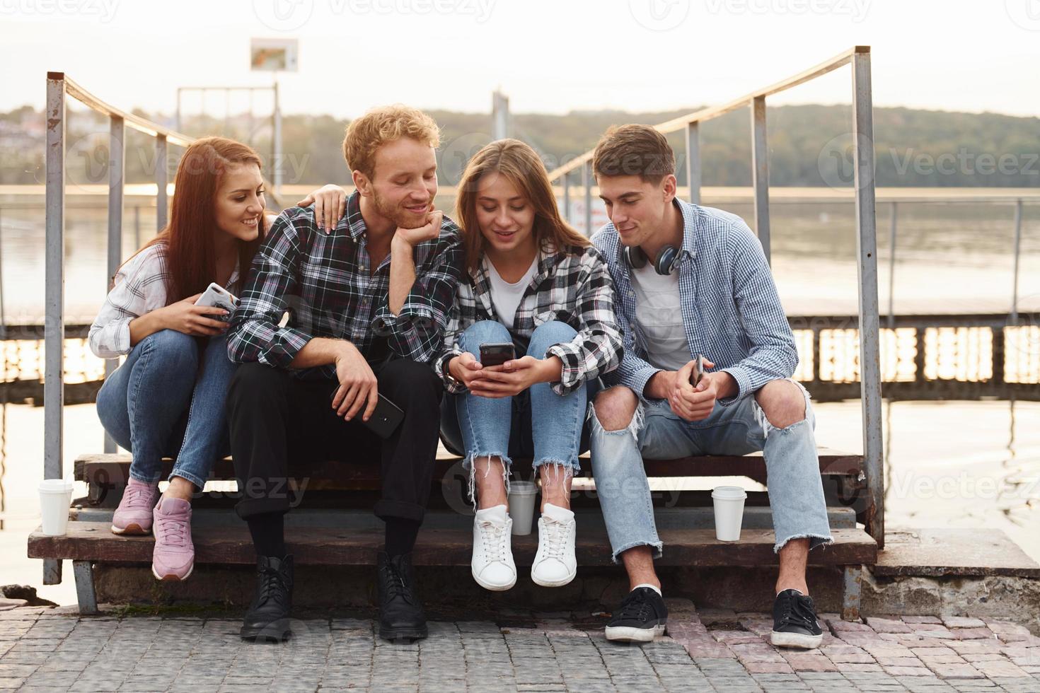 Sitting near lake and embracing each other. Group of young cheerful friends that is outdoors having fun together photo