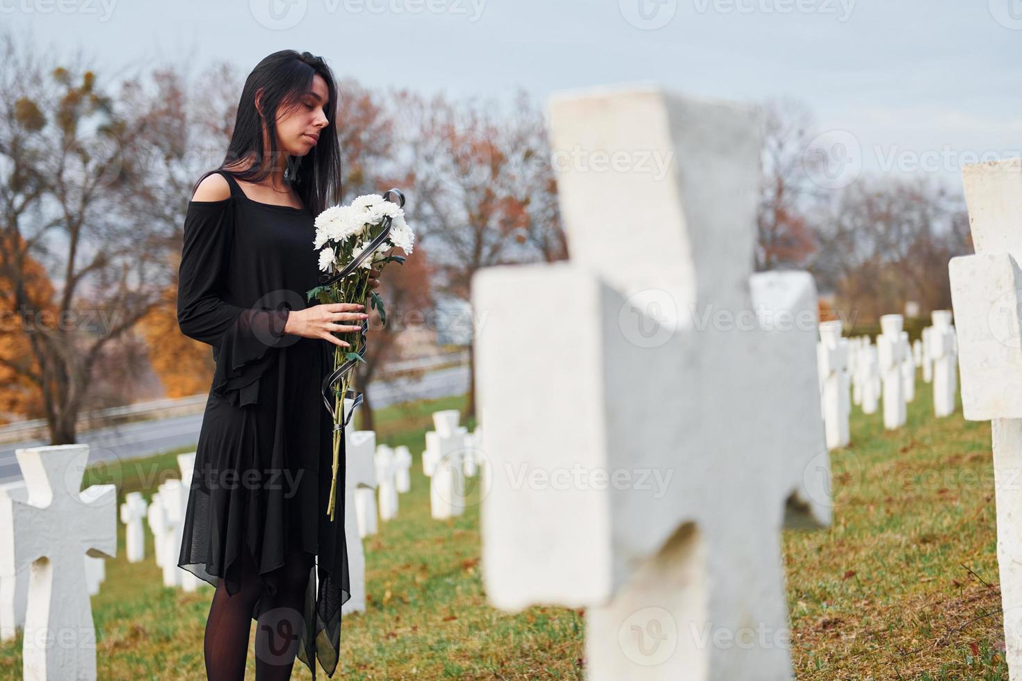 With flowers in hands. Young woman in black clothes visiting cemetery with many white crosses. Conception of funeral and death photo