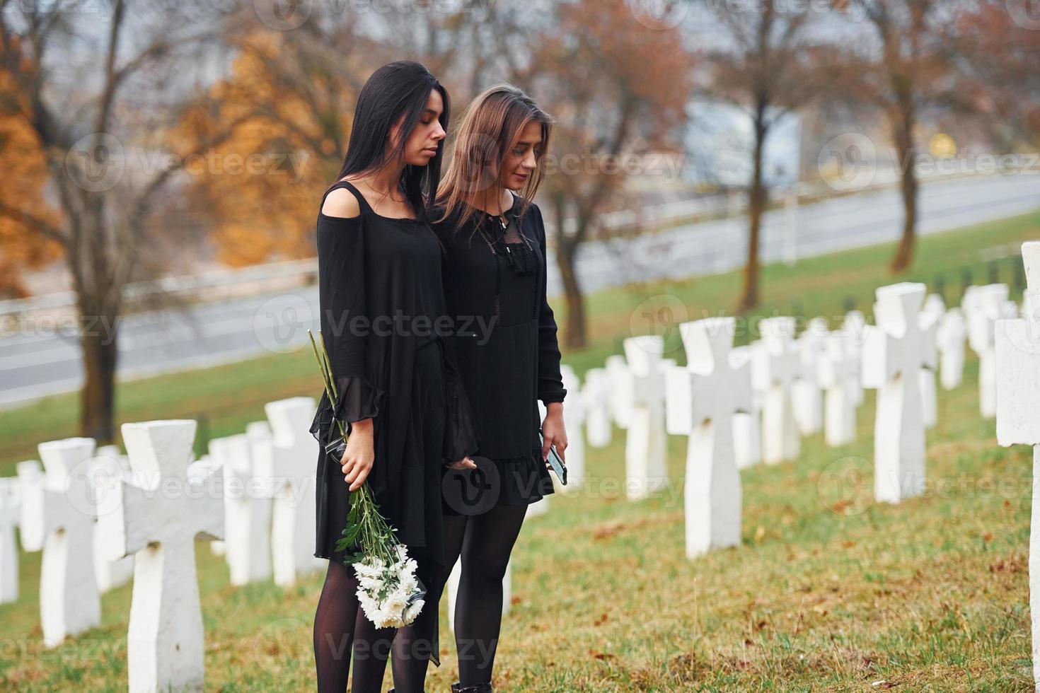 Holds flowers. Two young women in black clothes visiting cemetery with many white crosses. Conception of funeral and death photo